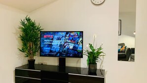 A living room with a flat-screen TV mounted on a black entertainment unit, flanked by two potted plants. A wall clock is visible above the TV.