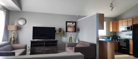 En moderne stue med et fladskærms-tv, sofaer, et sofabord og et spiseområde med et køkken i baggrunden.