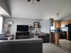 A modern living room with a flat-screen TV, sofas, a coffee table, and a dining area with a kitchen in the background. - Golden Horizon Home | Edmonton | 4 Bed • 2 Bath. 1 additional bed (Edmonton)