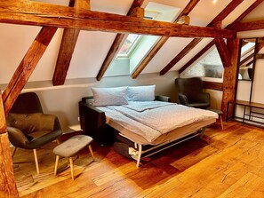 Une chambre avec un plafond en pente, des poutres en bois, un lit, une chaise et un petit tabouret.