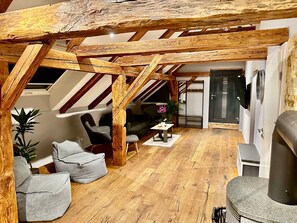 A loft-style room with exposed wooden beams, a sofa, chairs, a small table with a vase, and a kitchen area with a stove. - Apartment 'Ferienwohnung Weißensberg' with Balcony and Wi-Fi (Weißensberg)