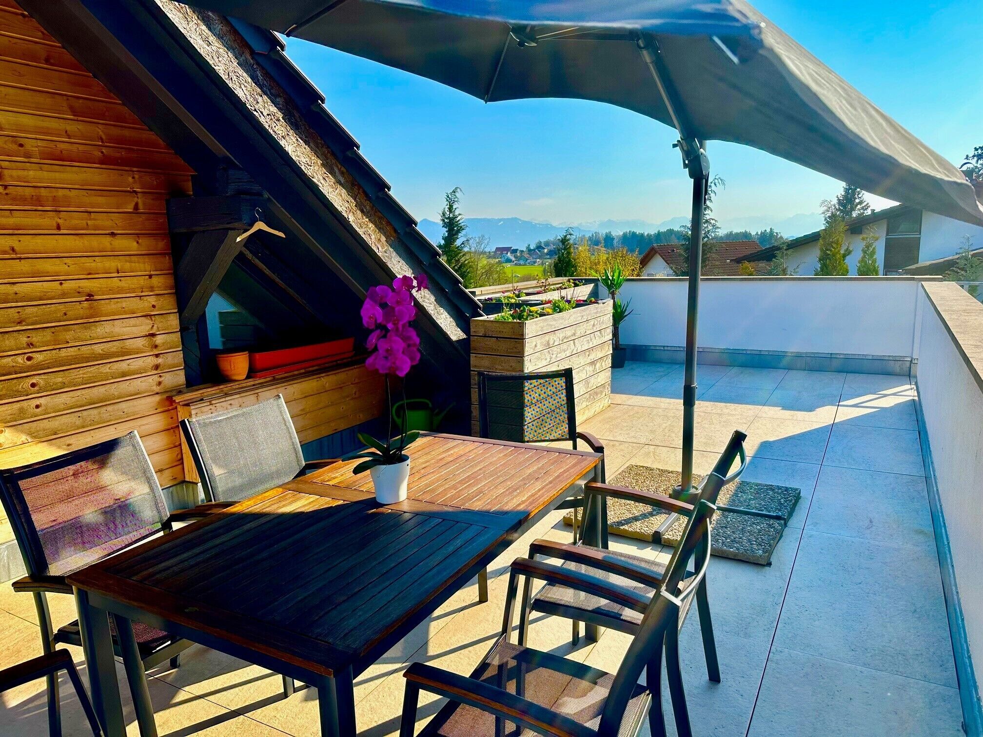 Eine Dachterrasse mit einem Holztisch, Stühlen, einer hängenden Blume und Blick auf die Berge.