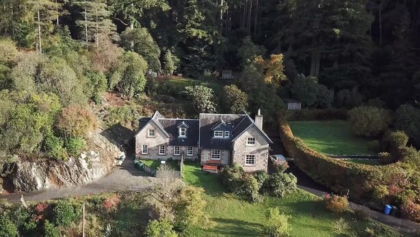 Une maison en pierre, dotée d’un toit à forte pente, entourée d’une végétation luxuriante et d’une pelouse bien entretenue.