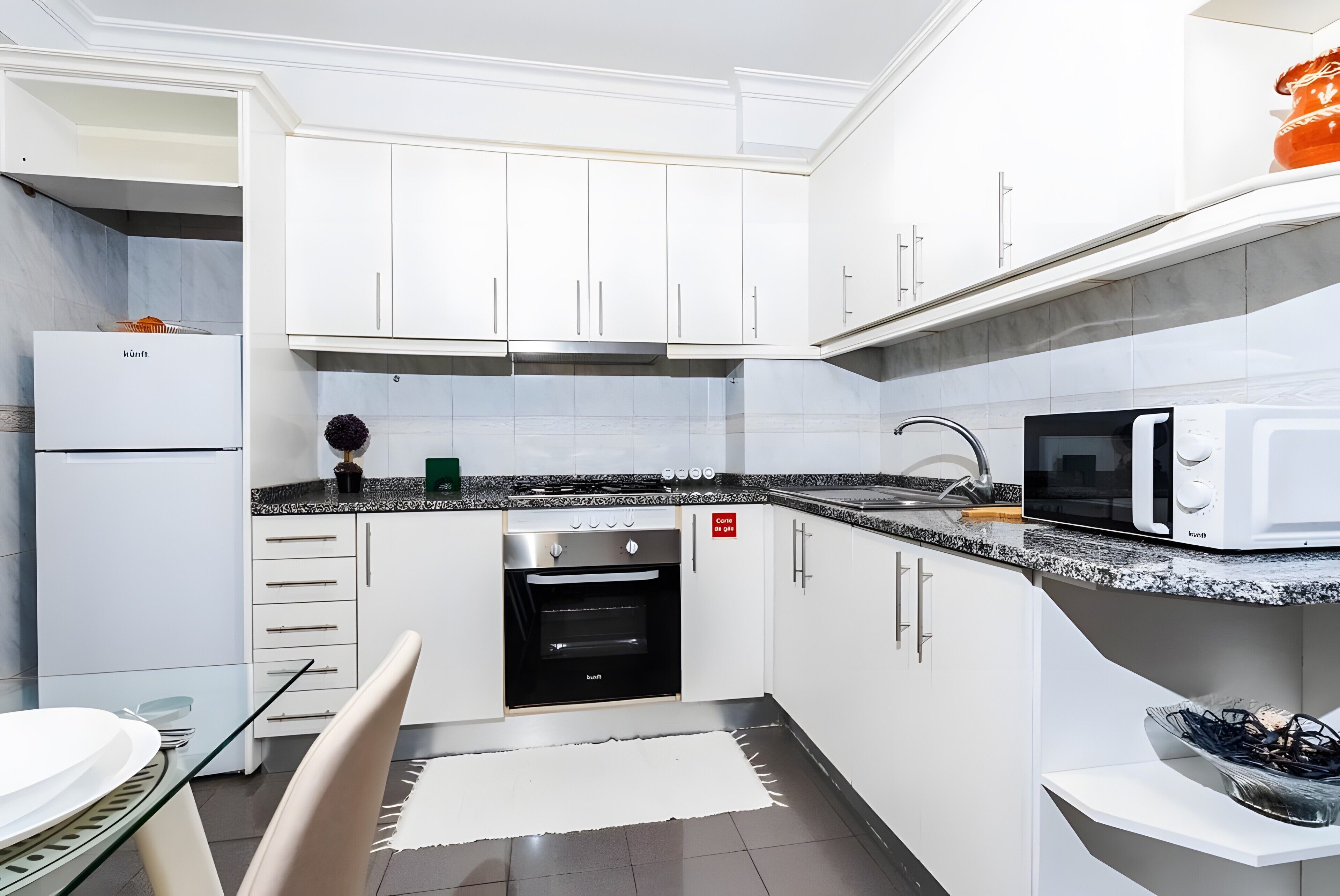 A modern kitchen with white cabinets, a built-in oven, a microwave, and a glass dining table.