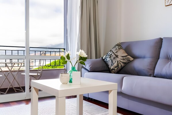 A modern living room with a grey sofa, a white side table, and a balcony with a view.