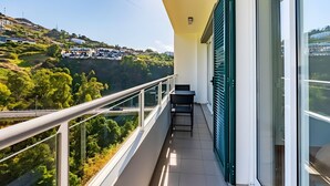 A balcony with a view of a hillside, a table, and a chair. - Casa Sol – Modern Apartment in Central Caniço with Pool and Parking (Caniço)