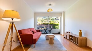 A modern living room with a red sofa, a tripod lamp, a round wooden side table, and a view of a green landscape through a large window. - Casa Sol – Modern Apartment in Central Caniço with Pool and Parking (Caniço)