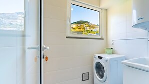 A laundry room with a washing machine, a dryer, a window with a view of a hillside, and a vent hood. - Casa Sol – Modern Apartment in Central Caniço with Pool and Parking (Caniço)