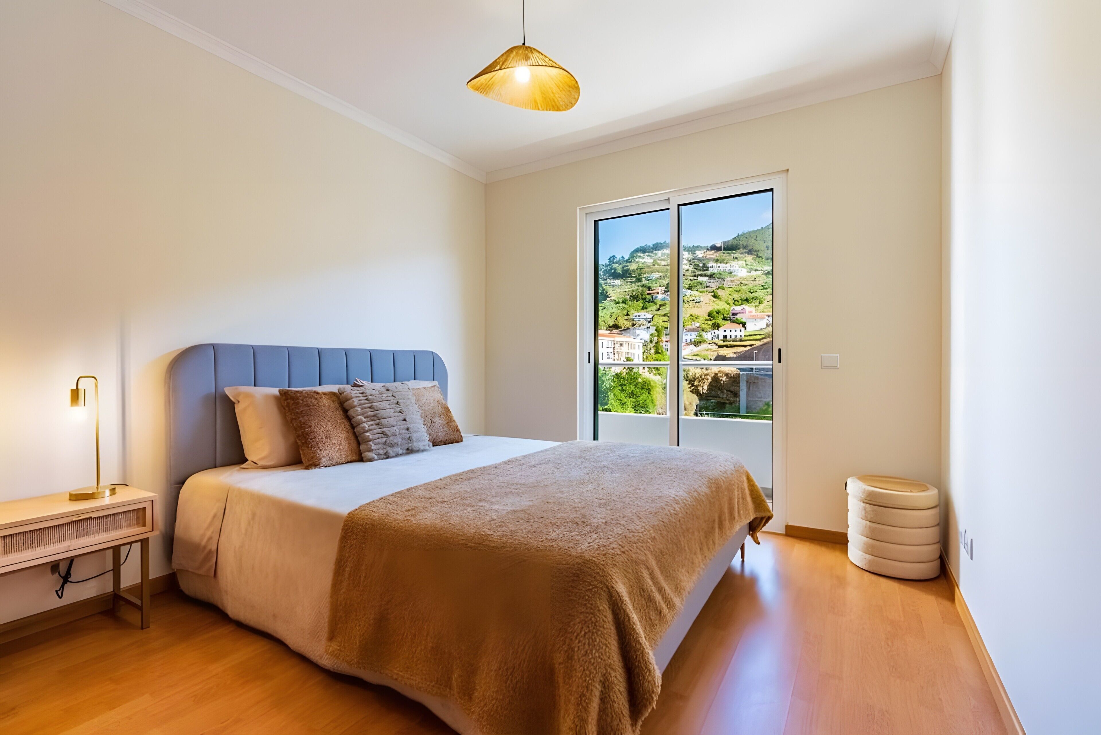 Een slaapkamer met een bed, een nachtkastje, een lamp, een raam met uitzicht op een heuvel, en een plafondlamp.