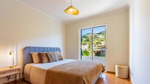 A bedroom with a bed, a bedside table, a lamp, a window with a view of a hillside, and a ceiling light. - Casa Sol – Modern Apartment in Central Caniço with Pool and Parking (Caniço)