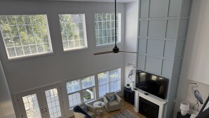 A spacious living room with a fireplace, large windows, and a high ceiling.