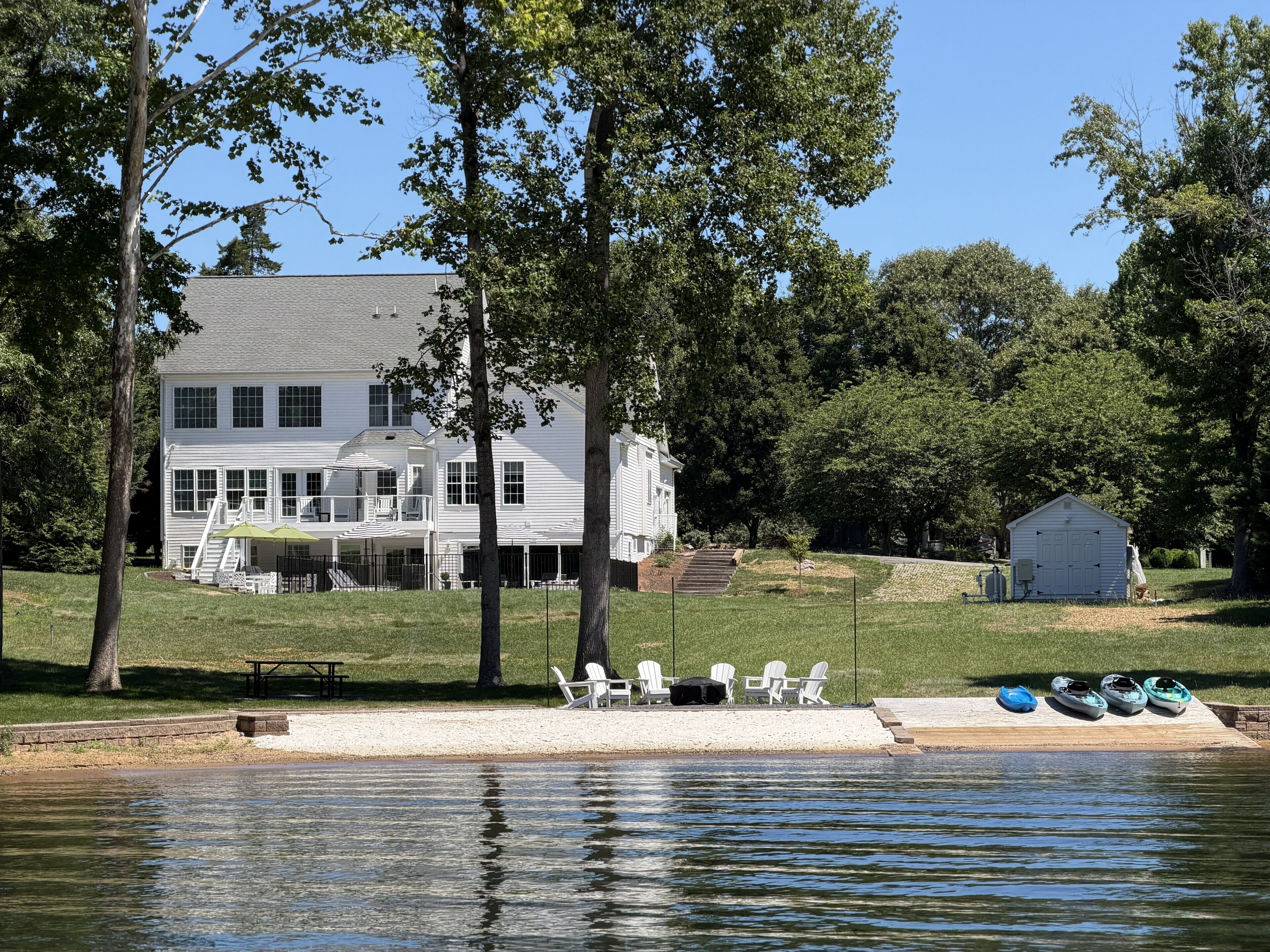 Una casa de dos pisos con un balcón, un muelle con sillas y kayaks, y una caseta pequeña.