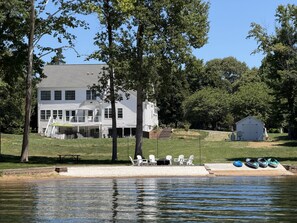 Une maison à deux étages avec un balcon, une jetée équipée de chaises et de kayaks, et un petit abri de jardin.