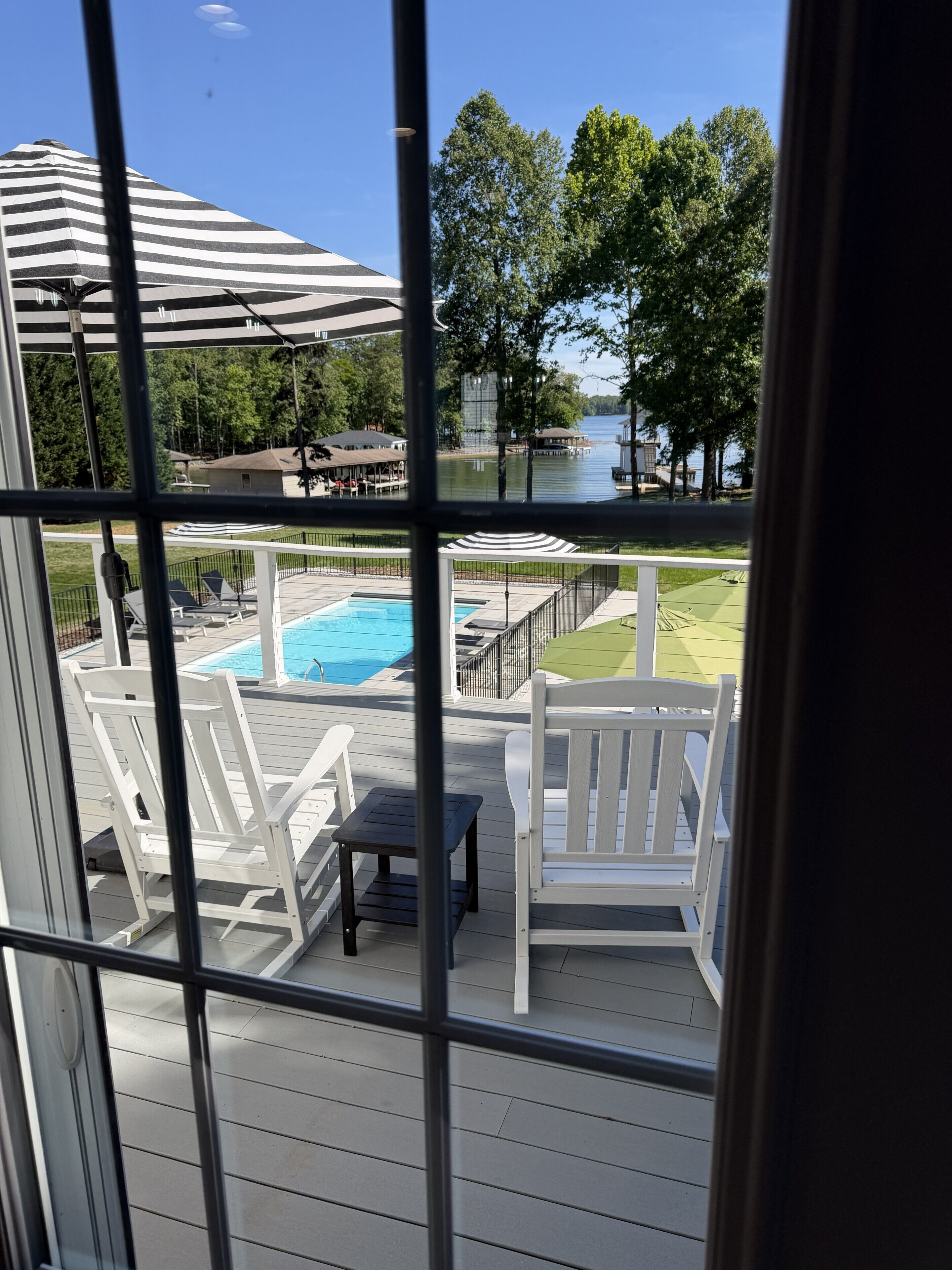 A view of a swimming pool area with lounge chairs and umbrellas, a dock extending into a body of water, and a grassy area with trees.