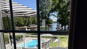 A view of a swimming pool area with lounge chairs and umbrellas, a dock extending into a body of water, and a grassy area with trees.