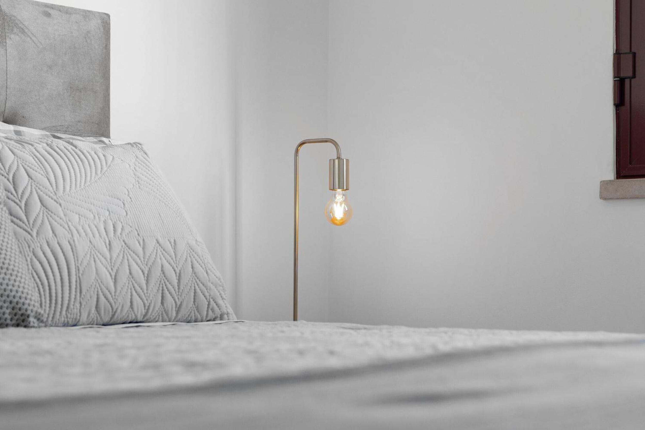 A bed with a textured white blanket and pillows, a modern floor lamp with a visible light bulb, and a plain wall with a small window.