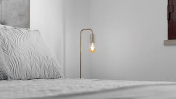 A bed with a textured white blanket and pillows, a modern floor lamp with a visible light bulb, and a plain wall with a small window.