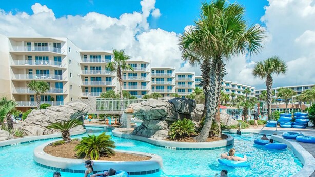 A swimming pool area with multiple levels of hotel buildings, palm trees, and people enjoying the water. - Waterscape 424-b (Fort Walton Beach)