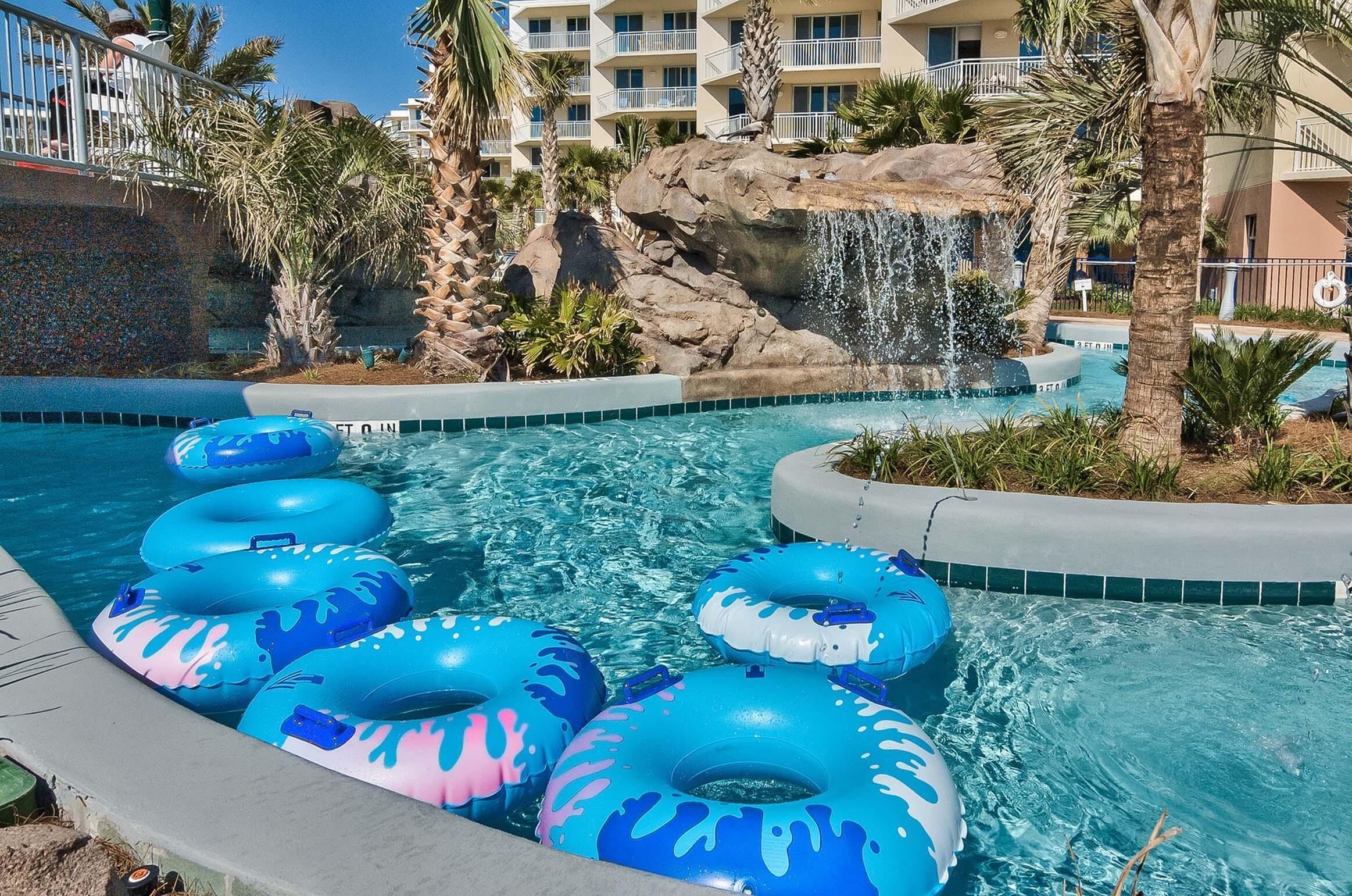 Una piscina con varios flotadores azules y rosas, una cascada, palmeras y un edificio con balcones.