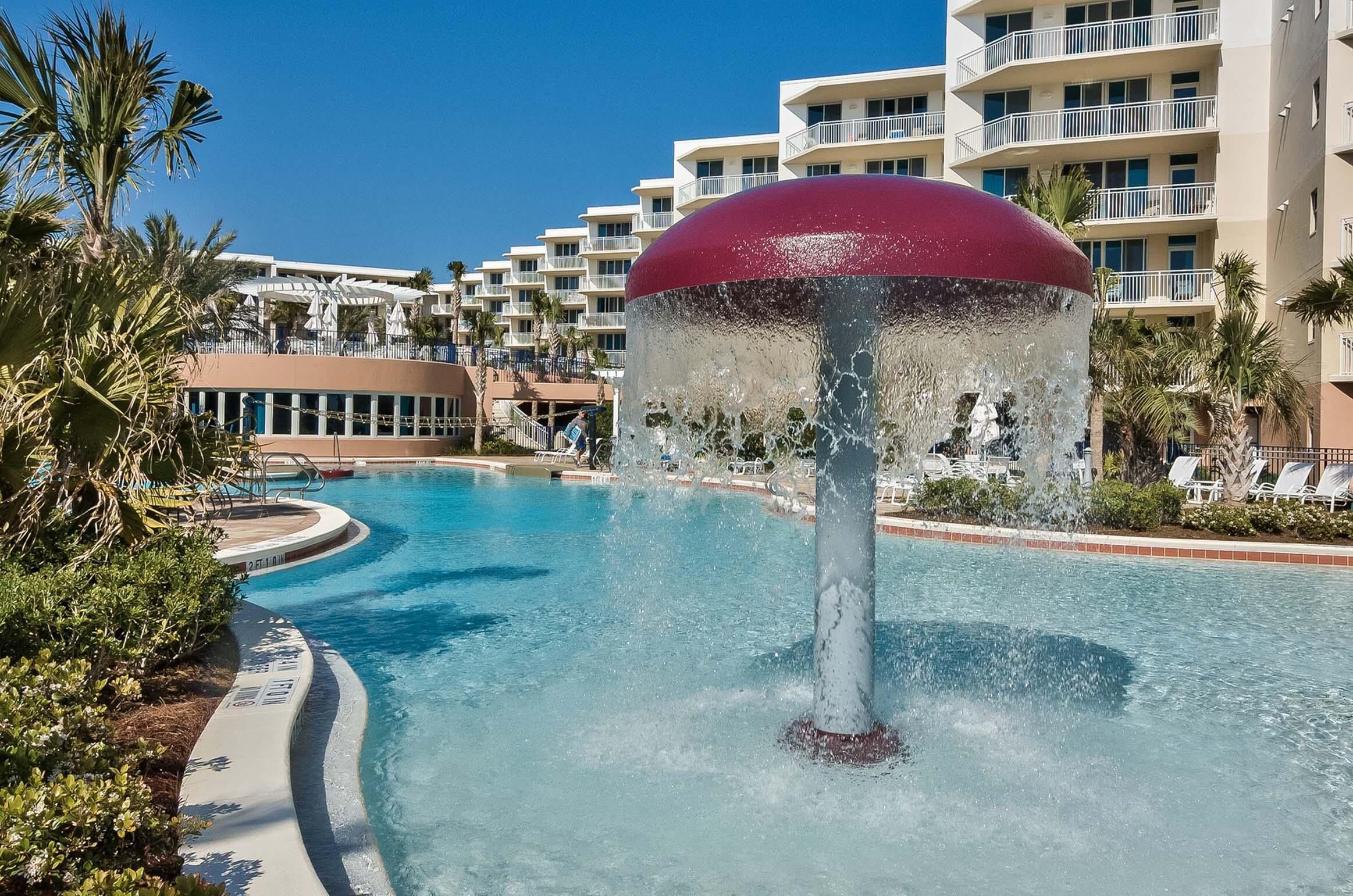 A swimming pool with a water fountain, palm trees, and a hotel building in the background.