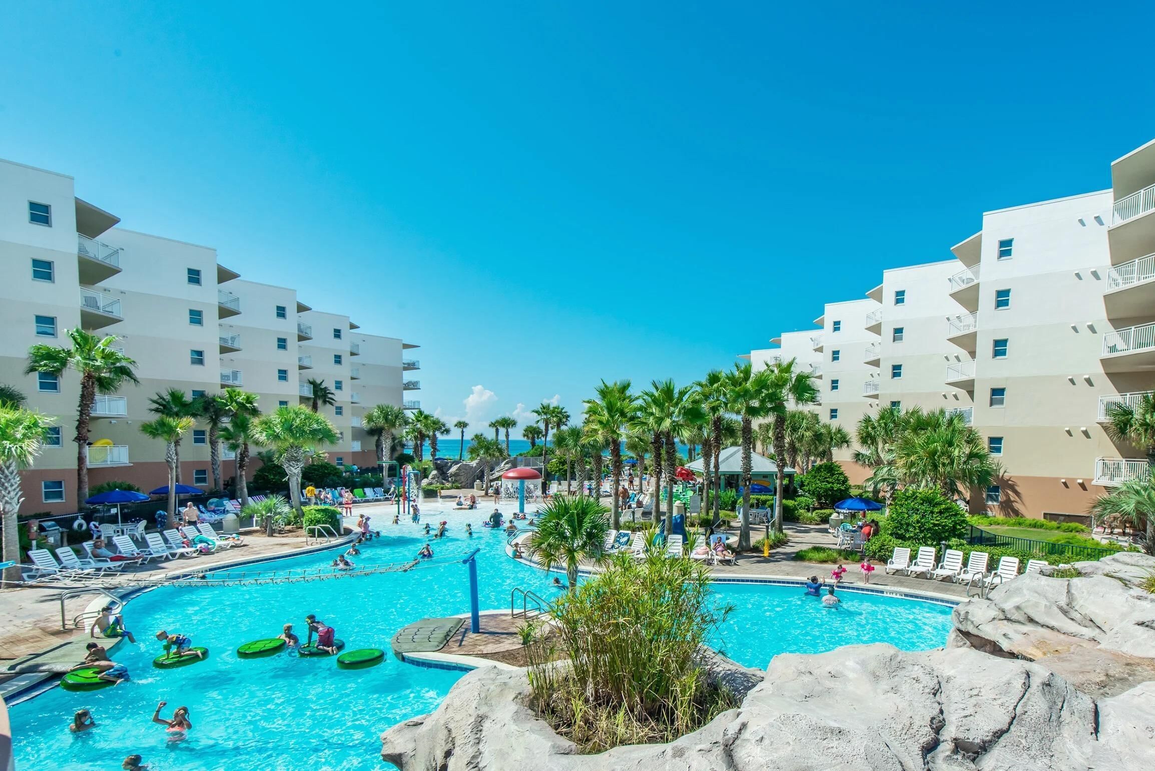 A resort pool area with multiple swimming pools, palm trees, and a building with balconies.