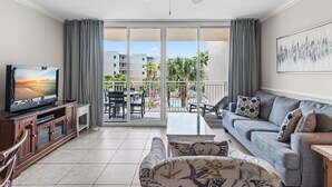 A living room with a TV, sofa, and a balcony with a pool view. - Waterscape 424-b (Fort Walton Beach)