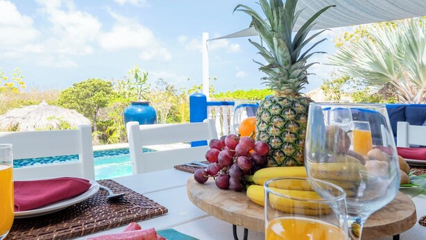 Une table dressée pour le petit-déjeuner, avec des fruits, du pain et des verres de jus, donnant sur une piscine et un espace verdoyant.