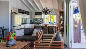 Un salon moderne avec un canapé, des tables en bois, des chaises en osier, une cuisine avec des meubles blancs et une grande fenêtre donnant sur un paysage verdoyant.