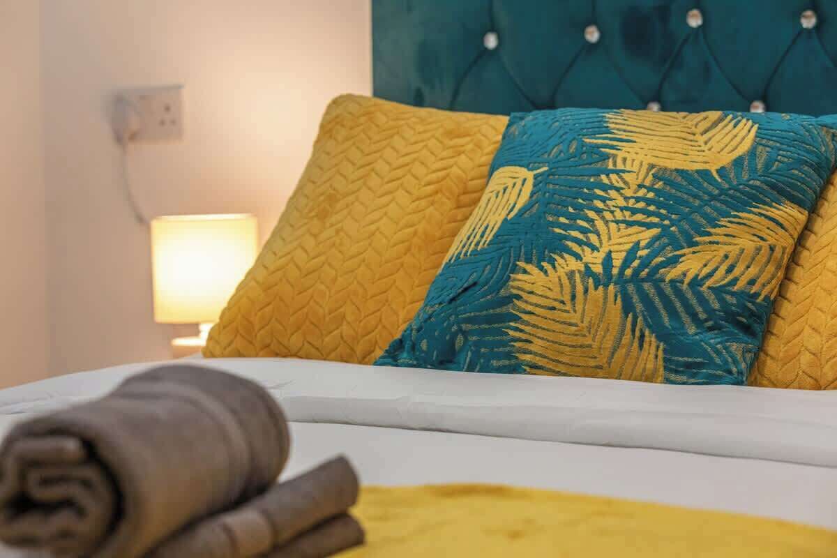 A bed with a white mattress, a rolled-up brown towel, and two decorative pillows with a leaf pattern.