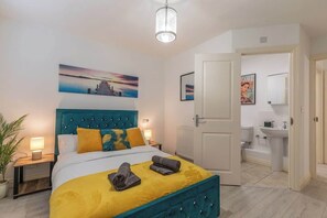 Ein Hotelzimmer mit einem Bett, einem Nachttisch, einer Topfpflanze, einem Gemälde, einem Badezimmer mit Waschbecken und Toilette sowie einer Tür zu einem anderen Raum.