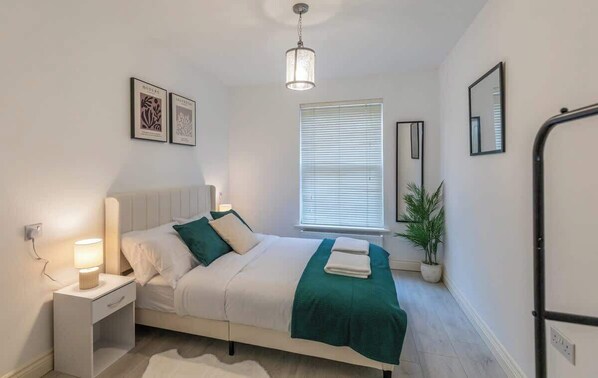 A modern bedroom with a bed, bedside lamp, window with blinds, and a potted plant.