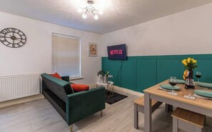 Ein modernes Wohnzimmer mit einem grünen Sofa, einem Esstisch gedeckt mit Weingläsern, einer Uhr und einem Fernseher, der 'NETFLIX' zeigt.