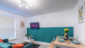 Uma sala de estar moderna com paredes verde-água, uma mesa de jantar de madeira posta com copos de vinho, uma pequena mesa redonda com uma planta e uma garrafa, e um sinal da Netflix fixado na parede.
