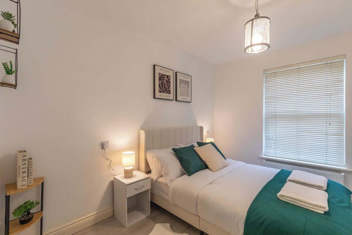 Une chambre à coucher comprenant un lit, des tables de chevet, une commode, une lampe, une fenêtre avec des stores et des œuvres d’art encadrées accrochées au mur.