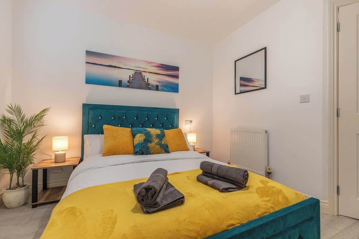 Una cama bien hecha con una colcha amarilla y dos toallas dobladas, un cabecero color turquesa y una pintura de atardecer arriba.