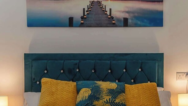Cama com cabeceira azul-turquesa, duas almofadas estampadas com folhas tropicais e duas toalhas dobradas sobre um edredom amarelo.