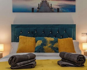 A bed with a teal headboard, two pillows with tropical leaf patterns, and two folded towels on a yellow bedspread.
