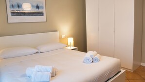 Ein ordentlich bezogenes Bett mit weißen Leinentüchern, einer Nachttischlampe und aufgerollten Handtüchern daneben.