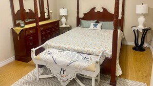 A four-poster bed with a floral bedspread, a wooden dresser with drawers, a mirror, a bedside table with a lamp, and a rocking chair with a decorative tablecloth. - Clean, Cozy and Spacious 1-Bedroom Apartment Near Airport and City Center (St. John's)