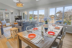 A dining area with a wooden table set for four, a grey sofa, and a TV in the background. - Peaceful home with dock, kayaks and sunset views (Deltaville)