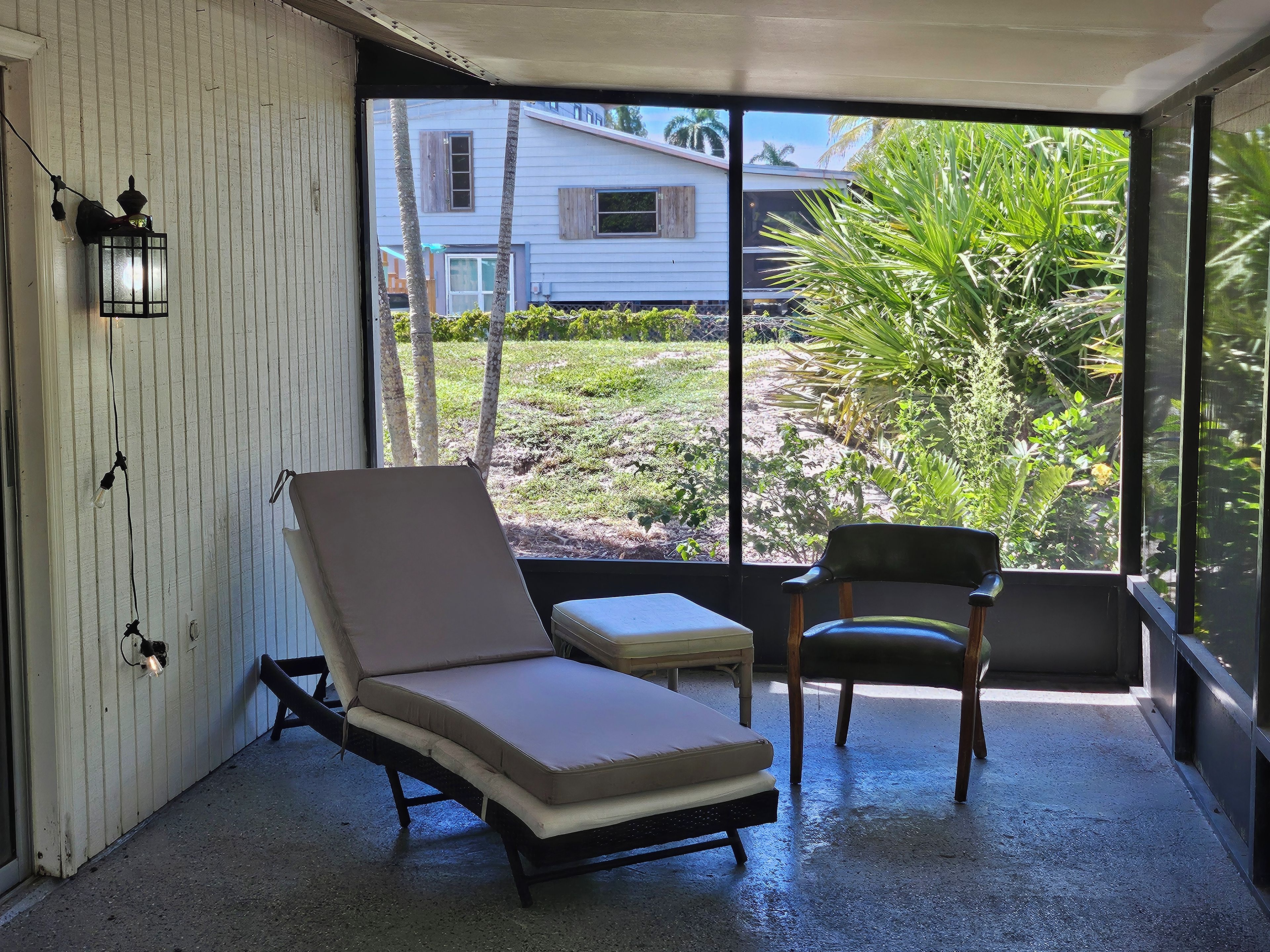 Une véranda avec vue sur une maison et des palmiers, comprenant un fauteuil de relaxation et une petite table.