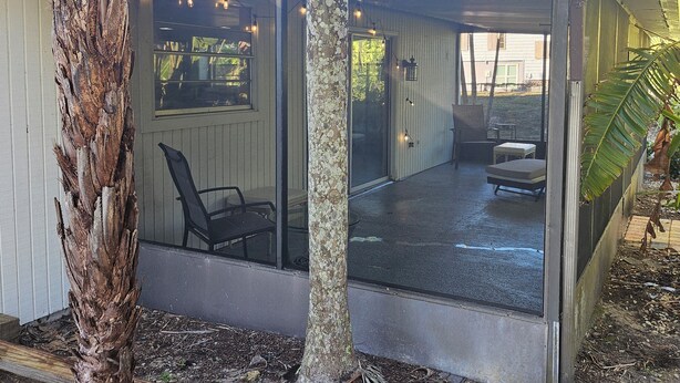 A covered outdoor seating area with a chair and a table, a glass door leading inside, and a tree in the foreground.