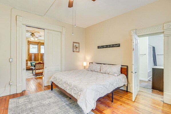 A bedroom with a large bed, a wooden floor, a ceiling fan, and a door leading to a bathroom.