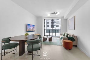 A modern living room with a dining area, a sofa, and a large window with a city view. - Stylish High-Rise Living | Pool, Spa & Cowork (Miami)
