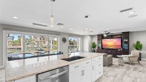 Une cuisine moderne dotée d’un grand îlot central, d’un coin repas avec télévision et offrant une vue sur une piscine et des bâtiments.