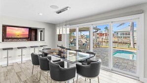 Un espace repas moderne avec une table en verre et des chaises noires, un grand téléviseur fixé au mur et une vue sur la piscine à travers des portes vitrées.
