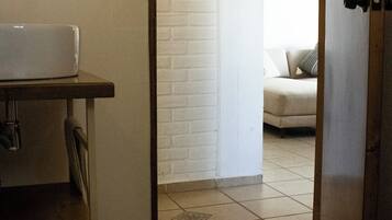 A bathroom with a toilet, a sink, and a white brick wall. There is a door leading to a living area with a couch and a painting.
