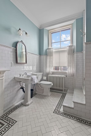 A bathroom with white tiled walls, a white sink, a toilet, and a bathtub. There is a window with curtains and a mirror above the sink. - CIRCA 1800 TWO-STORY EXECUTIVE RENTAL SUITE (St. John's)