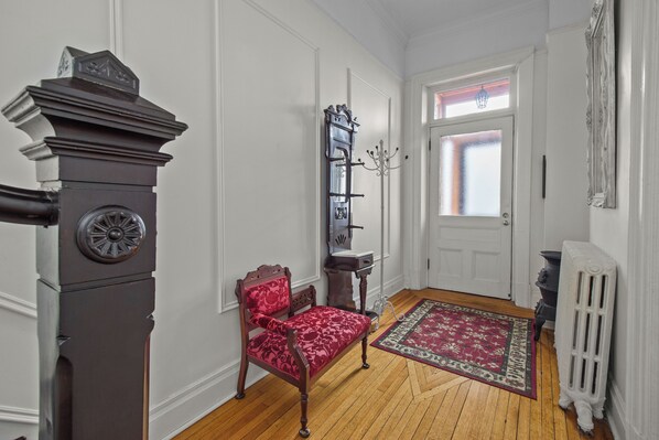 A hallway with a wooden floor, a red upholstered chair, a white door with a frosted glass panel, a radiator, and a wall-mounted coat rack. - CIRCA 1800 TWO-STORY EXECUTIVE RENTAL SUITE (St. John's)