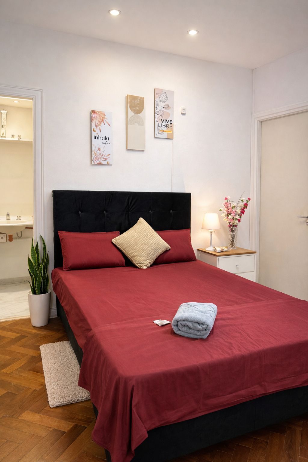 A neatly arranged bedroom with a bed, a bedside table with a lamp, a potted plant, and wall art.